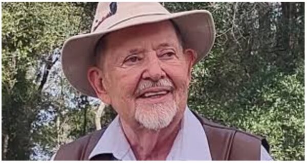 An elderly man with a white beard and mustache smiles while wearing a beige sun hat and a light blue shirt outdoors, surrounded by trees and greenery.