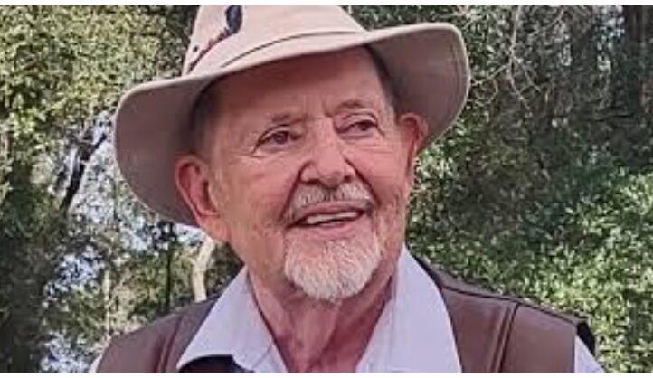 An elderly man with a white beard and mustache smiles while wearing a beige sun hat and a light blue shirt outdoors, surrounded by trees and greenery.