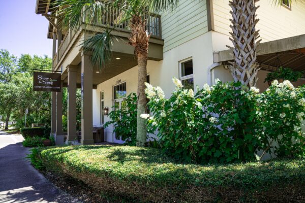A beige building with a covered entrance, palm trees, lush green bushes, and white flowers in front. A sign reads Song and Stone: Coffee, Sweets & Gelato. Sunlight and greenery surround the area.