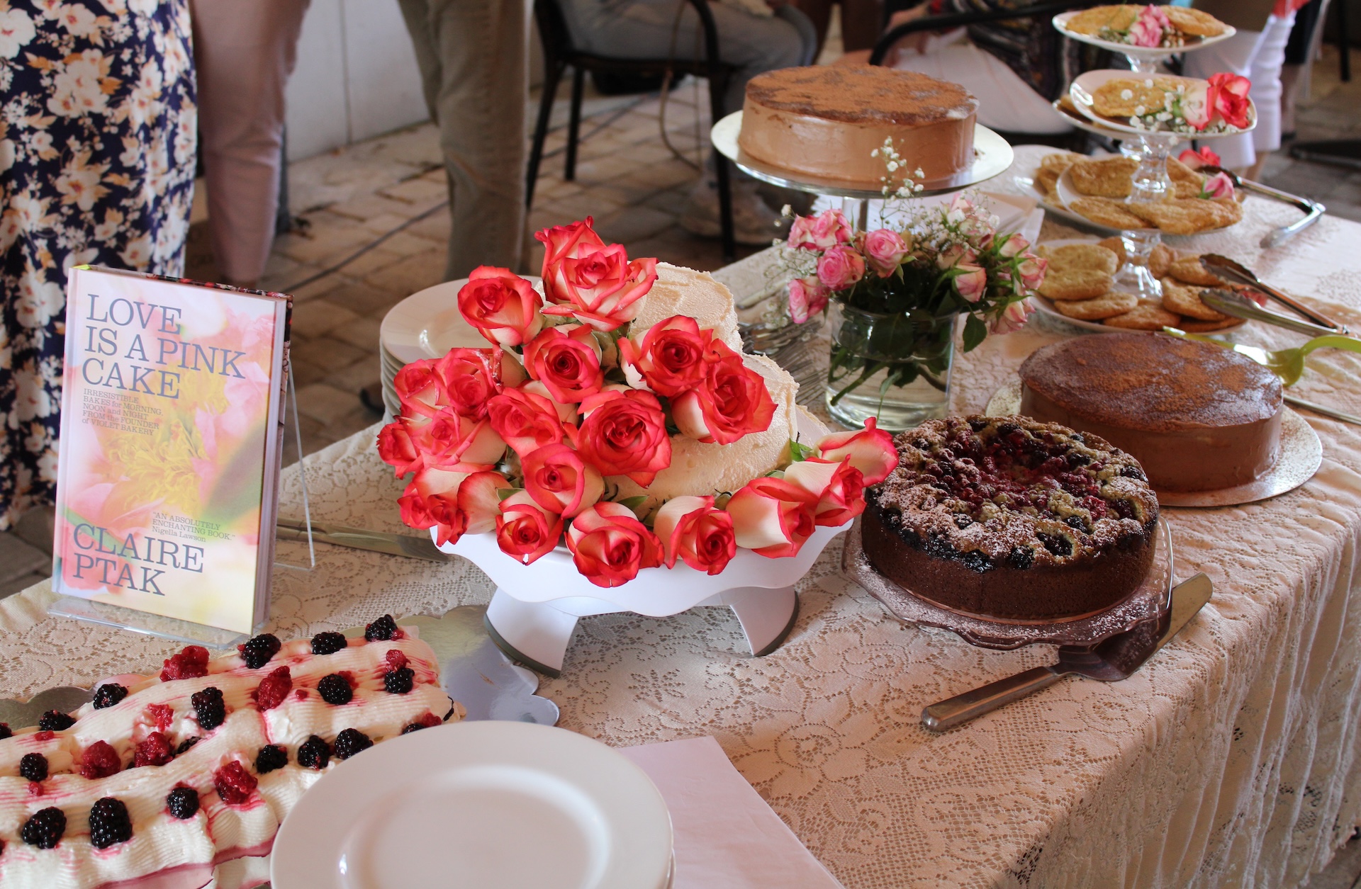 A table covered with a lace cloth displays various cakes, cookies, a plate of blackberries, a bouquet of pink roses, and a book titled LOVE IS A PINK CAKE by Claire Ptak.