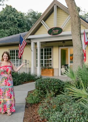 A woman in a floral dress stands in front of a yellow building with a sign that reads Sues Market and gestures toward the entrance.
