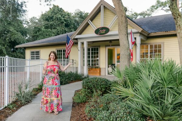 A woman in a floral dress stands in front of a yellow building with a sign that reads Sues Market and gestures toward the entrance.