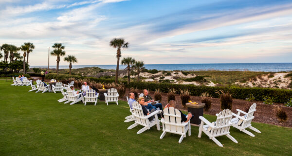 People sit in white Adirondack chairs on a grassy lawn near the beach, facing the ocean. Palm trees and sand dunes are in the background under a blue sky with wispy clouds.