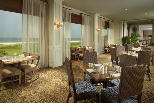 A bright restaurant dining area with wooden tables and wicker chairs, set with white dishes and yellow accents. Large windows offer views of dunes and the ocean outside. Light curtains and patterned carpet complete the elegant decor.