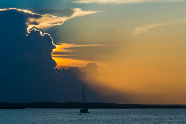 A sailboat floats on calm water at sunset, with dramatic clouds partially obscuring the sun, casting golden and blue hues across the sky and water.