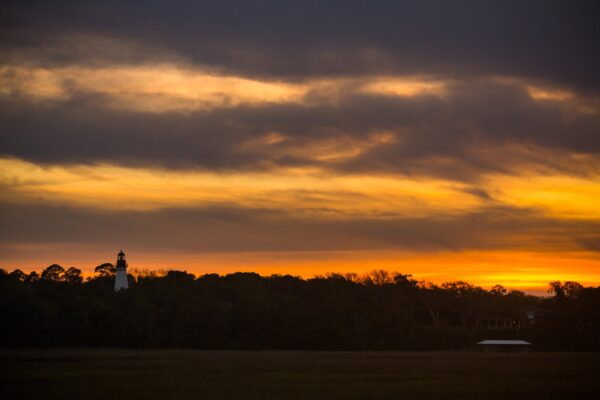 A vibrant orange and yellow sunset sky with dark clouds over a silhouette of trees. A small lighthouse and a building are visible among the trees on the horizon.