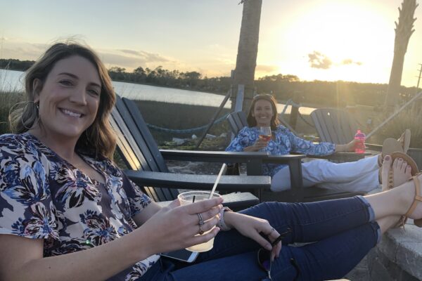 Two women relax in outdoor chairs by a lakeside at sunset, smiling and holding drinks. One has her legs up on a chair, and both are dressed in casual, patterned shirts. A hammock and water are visible in the background.