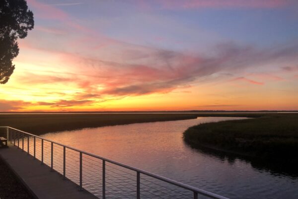 A winding river bordered by grass flows under a vivid sunset sky, with streaks of pink, orange, and yellow; a railing lines the riverbank in the foreground.