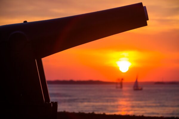 A cannon is silhouetted against a vibrant sunset over the water, with a sailboat and a small structure visible in the distance.