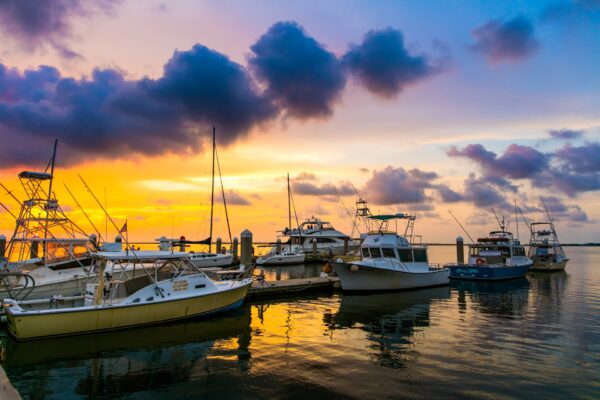 Several boats are docked at a marina during sunset, with vibrant orange and purple clouds reflecting on the calm water. The sky creates a dramatic, colorful backdrop for the peaceful scene.