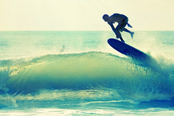 A surfer is airborne above a wave, balancing on a surfboard with the ocean and sunlight in the background. The water splashes up, and the scene captures the energy and motion of surfing.