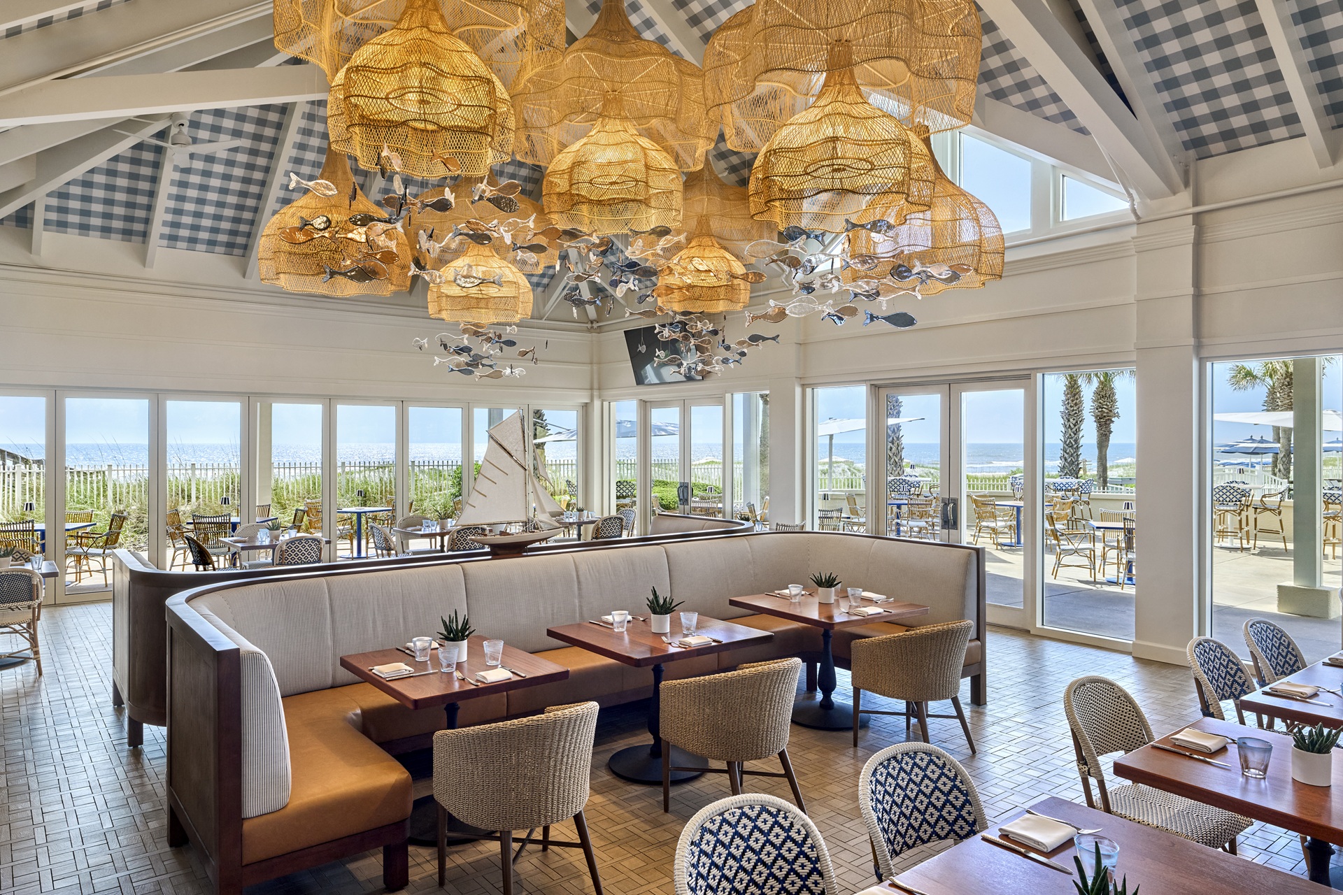 Bright coastal restaurant with wicker light fixtures, wood tables, cushioned chairs, and large windows showing an outdoor patio and an ocean view. Nautical decor and potted plants add to the airy, beachside atmosphere.