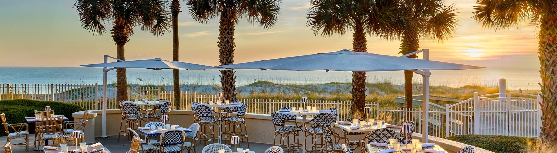 Outdoor restaurant with patterned chairs and tables on a patio, shaded by umbrellas. Palm trees line the space, and the sun is setting over the ocean in the background, creating a warm, inviting atmosphere.