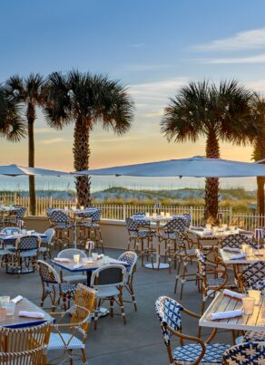 Outdoor restaurant with patterned chairs and tables on a patio, shaded by umbrellas. Palm trees line the space, and the sun is setting over the ocean in the background, creating a warm, inviting atmosphere.
