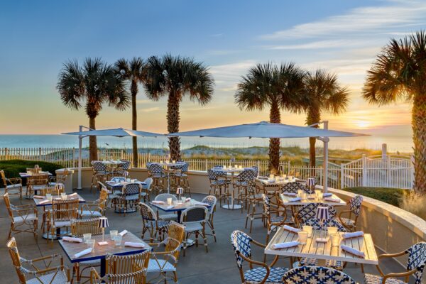 Outdoor restaurant with patterned chairs and tables on a patio, shaded by umbrellas. Palm trees line the space, and the sun is setting over the ocean in the background, creating a warm, inviting atmosphere.