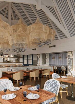 Coastal-style restaurant with high, beamed ceiling, wicker chandeliers, patterned blue and white chairs, wooden tables set with napkins and glasses, and a bright, airy ambiance.
