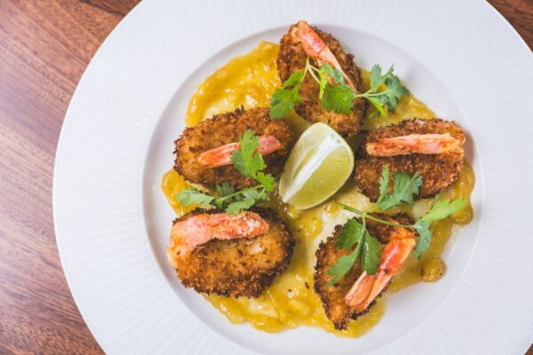 Five breaded shrimp pieces are arranged on a white plate with yellow sauce, garnished with fresh cilantro and a lime wedge, set on a wooden surface.