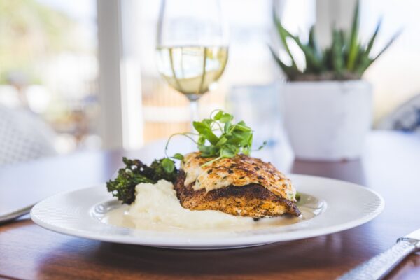 A plate with grilled fish atop mashed potatoes, garnished with greens and a side of broccoli, sits on a wooden table. A glass of white wine and a potted plant are in the background.