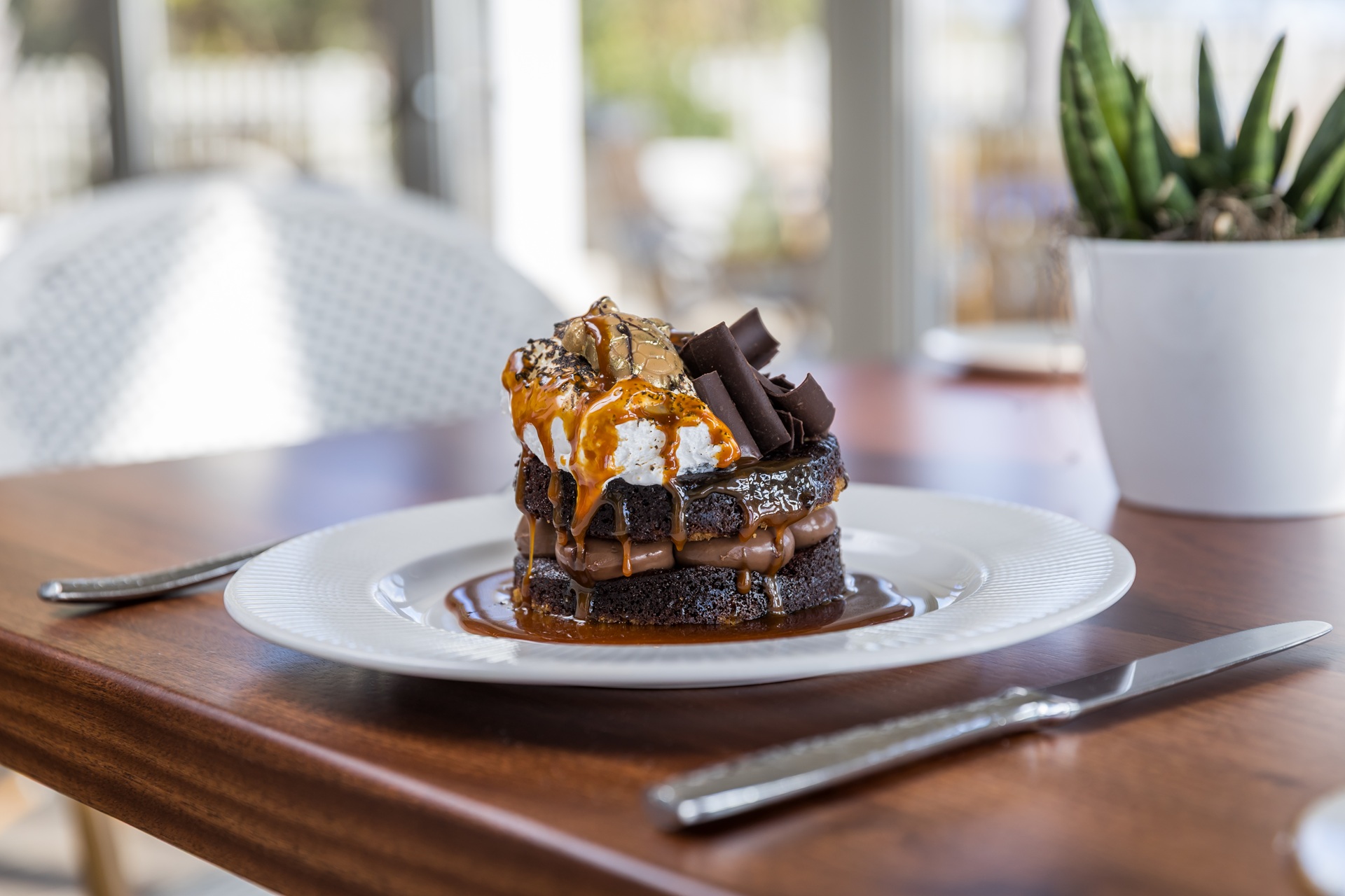 A decadent chocolate dessert with caramel sauce, whipped cream, and chocolate curls is served on a white plate beside a fork and knife on a wooden table, with a plant in the background.