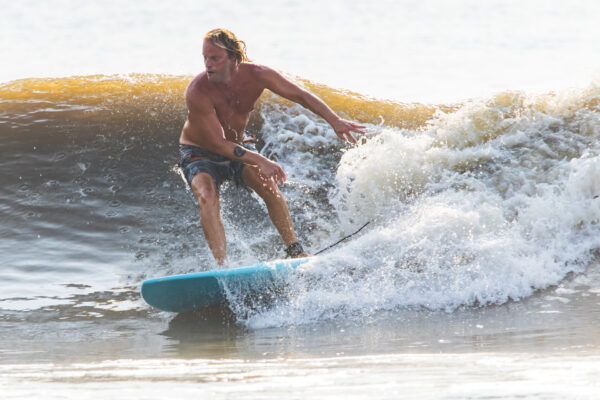 A man with long hair, wearing swim trunks and a smartwatch, surfs on a blue surfboard, riding a wave near the shore with water splashing around him.