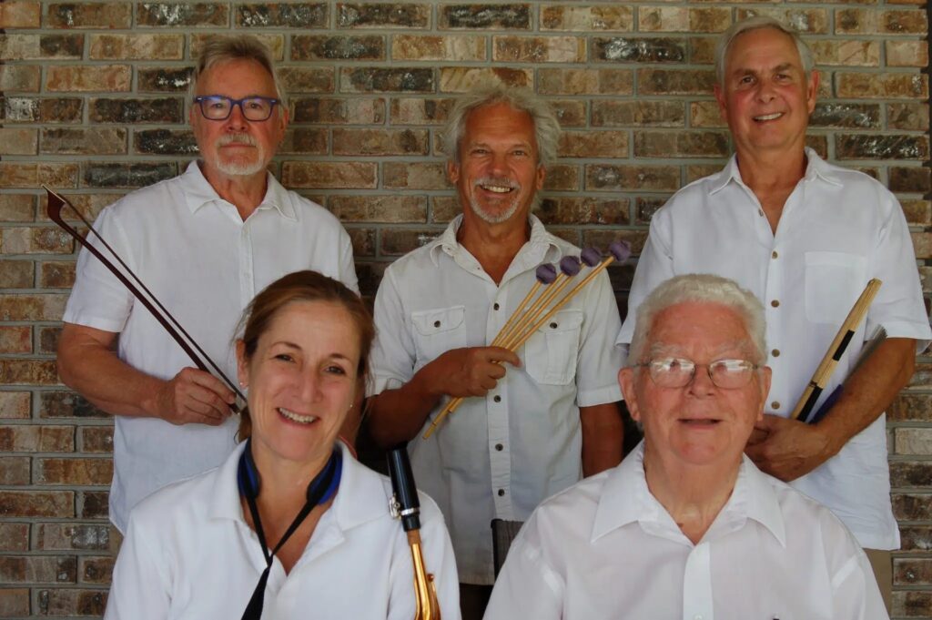 Five adults in white shirts pose in front of a brick wall, smiling. Three hold musical instruments: drumsticks, mallets, and a reed instrument. Two are seated in front; three stand behind them.