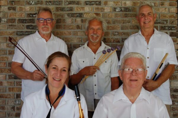 Five adults in white shirts pose in front of a brick wall, smiling. Three hold musical instruments: drumsticks, mallets, and a reed instrument. Two are seated in front; three stand behind them.