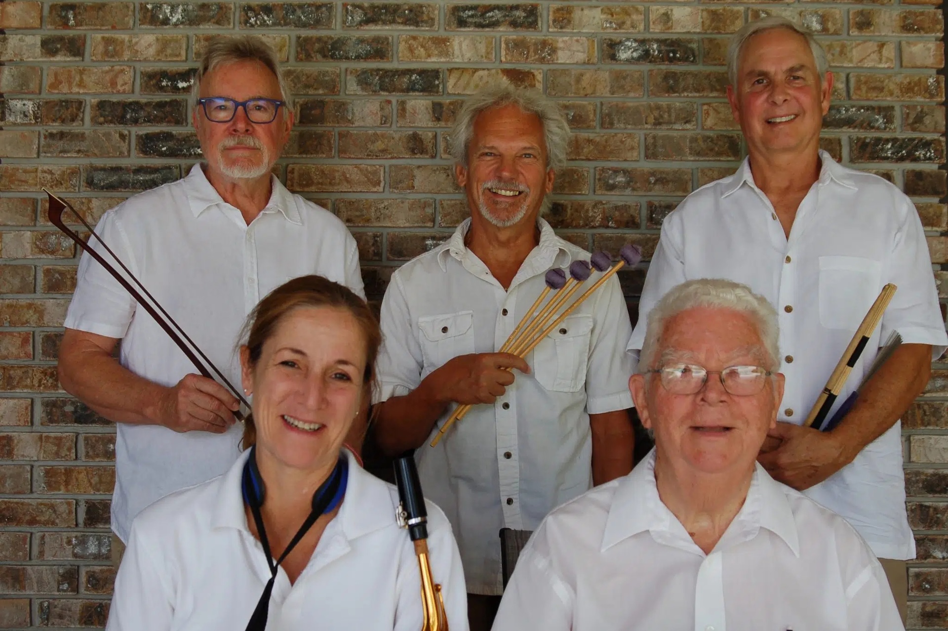 Five adults in white shirts pose in front of a brick wall, smiling. Three hold musical instruments: drumsticks, mallets, and a reed instrument. Two are seated in front; three stand behind them.