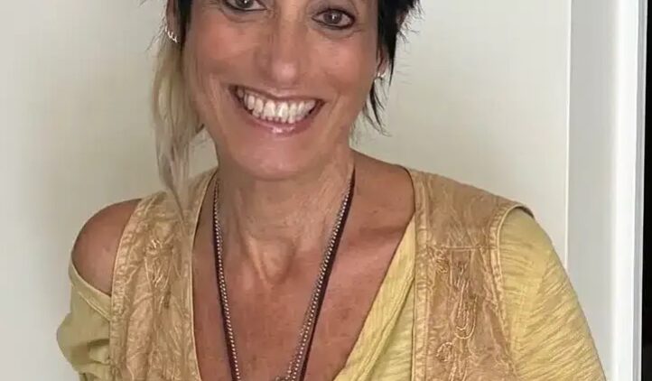 A smiling woman with short, spiky dark hair and a light streak stands against a white wall. She wears a yellow top, a beige vest, and layered necklaces, including one with a turquoise pendant.