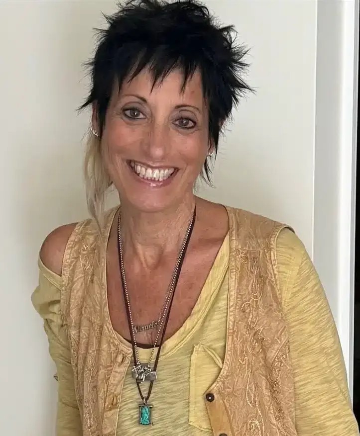 A smiling woman with short, spiky dark hair and a light streak stands against a white wall. She wears a yellow top, a beige vest, and layered necklaces, including one with a turquoise pendant.
