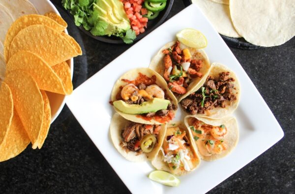 A square white plate with four assorted tacos, each topped with different meats, shrimp, avocado, and garnishes. Lime wedges are on the plate. Nearby are taco shells, tortillas, avocado, cilantro, jalapeños, and diced tomatoes.