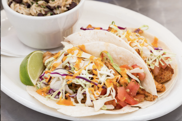 Two soft shell tacos filled with grilled chicken, shredded cabbage, diced tomatoes, and drizzled with orange sauce, served with a lime wedge. A bowl of rice and black beans is in the background.