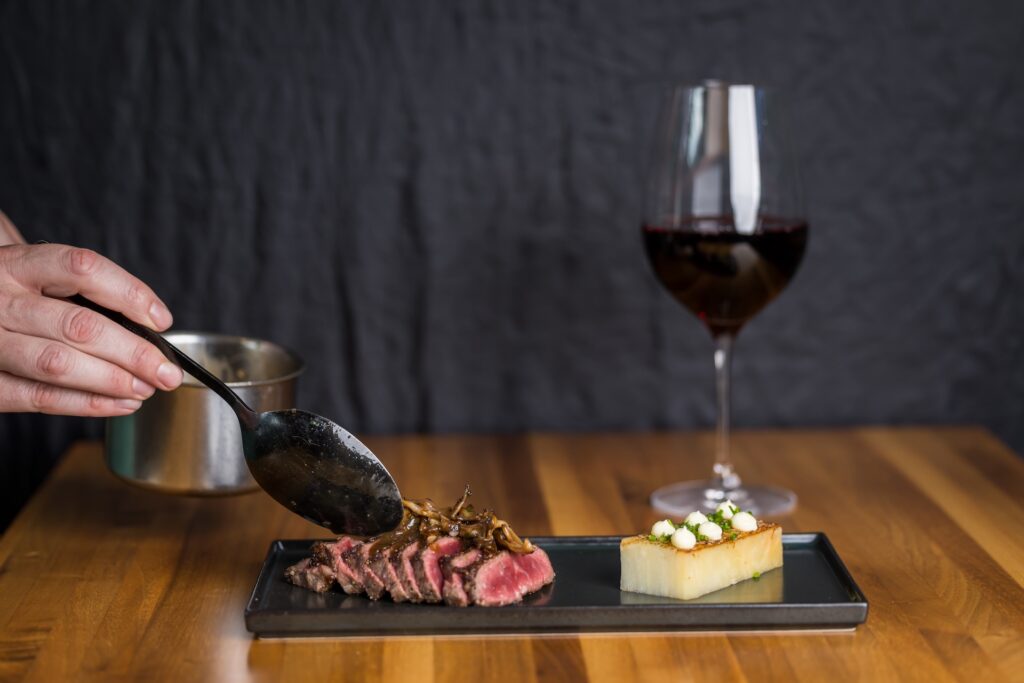 A person spoons sauce over sliced steak with mushrooms, served on a black plate with layered potatoes and a glass of red wine on a wooden table.