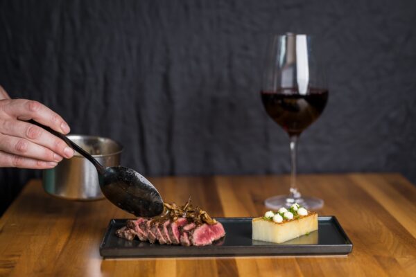 A person spoons sauce over sliced steak with mushrooms, served on a black plate with layered potatoes and a glass of red wine on a wooden table.