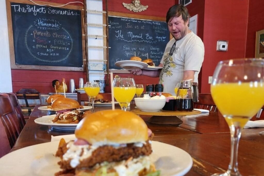 A server holding a plate of sliders walks past a long table set with multiple plates of sandwiches and glasses of orange juice, in front of two chalkboards listing menu items and bar information.