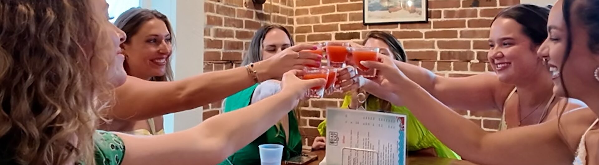 Six women at a restaurant table toast with small glasses of reddish drinks.