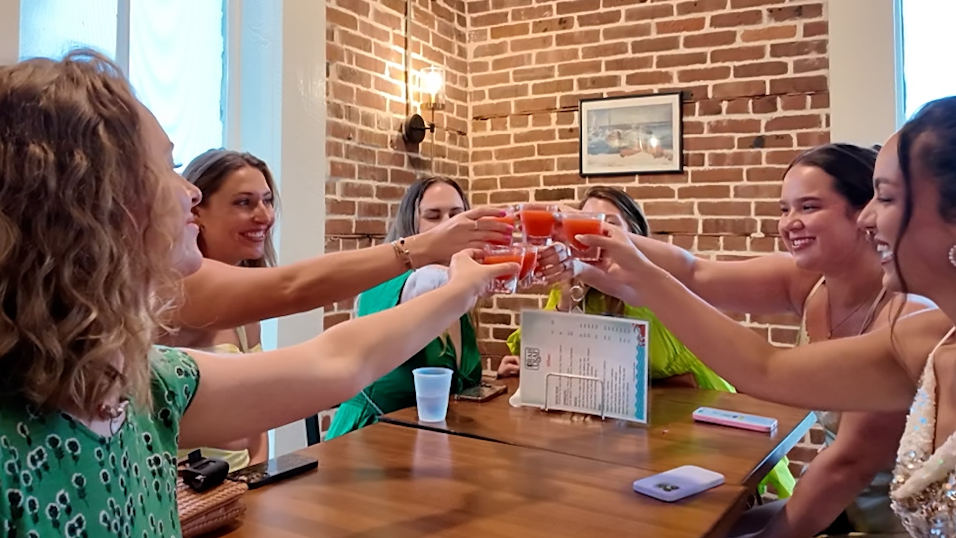 Six women at a restaurant table toast with small glasses of reddish drinks.