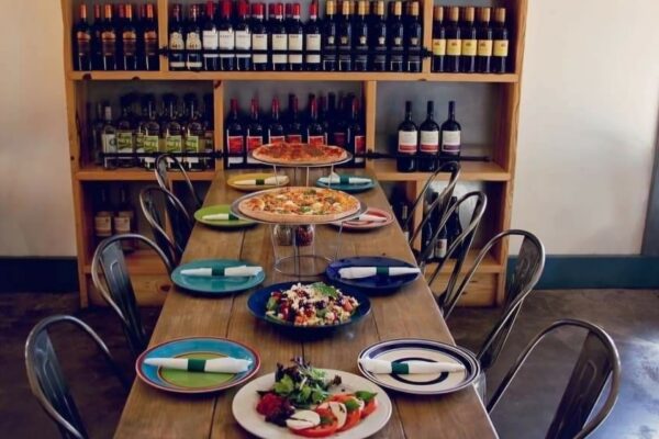 A long wooden dining table set for a meal with two pizzas on elevated stands, a salad, and a caprese salad, positioned in front of shelves filled with wine bottles.