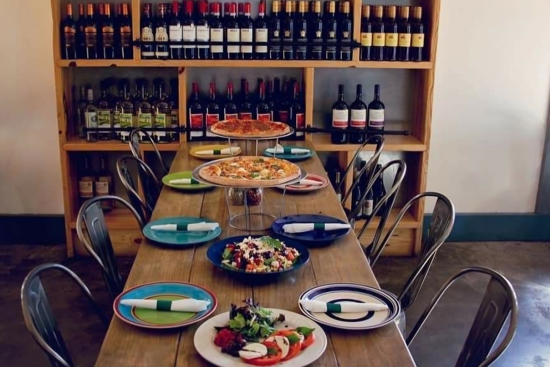 A long wooden dining table set for a meal with two pizzas on elevated stands, a salad, and a caprese salad, positioned in front of shelves filled with wine bottles.