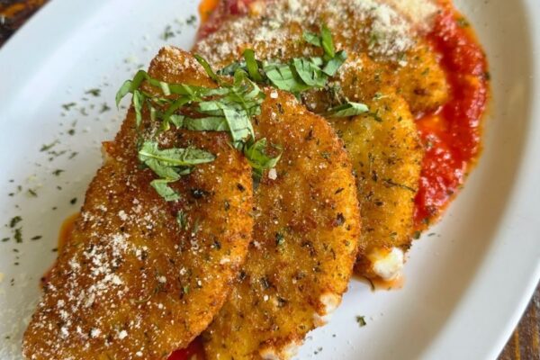 Three breaded, fried eggplant slices topped with grated cheese and fresh basil, served over a layer of tomato sauce on a white plate.