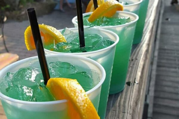 Three plastic cups filled with bright blue cocktails, ice, black straws, and orange slices, sitting on a wooden railing.