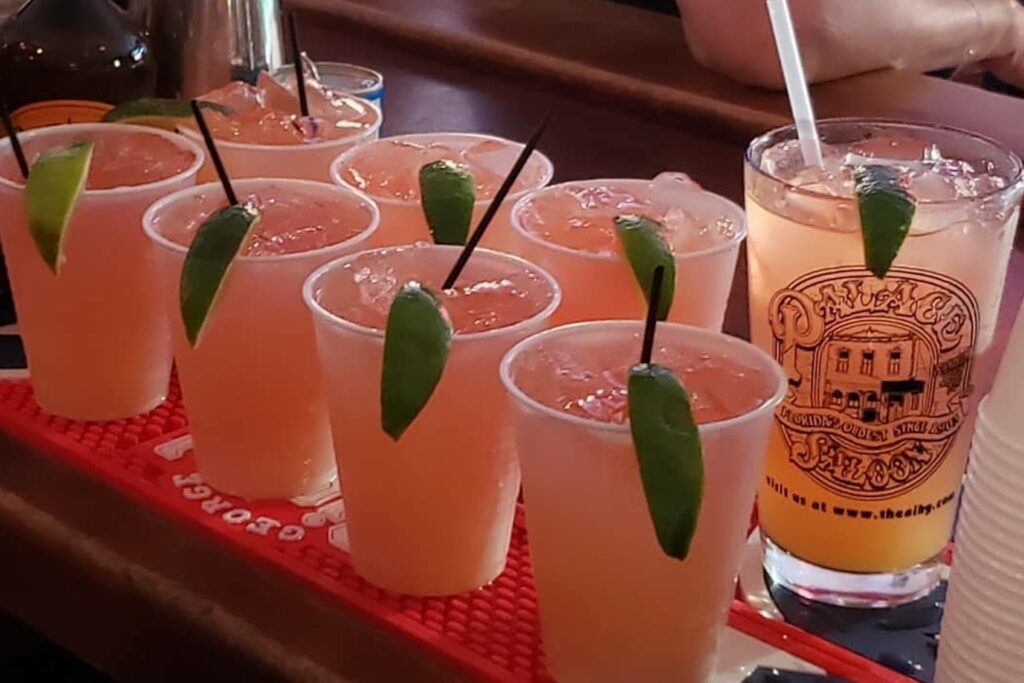 Several clear plastic cups filled with pink cocktails garnished with lime wedges, sitting on a red bar mat next to a branded glass.