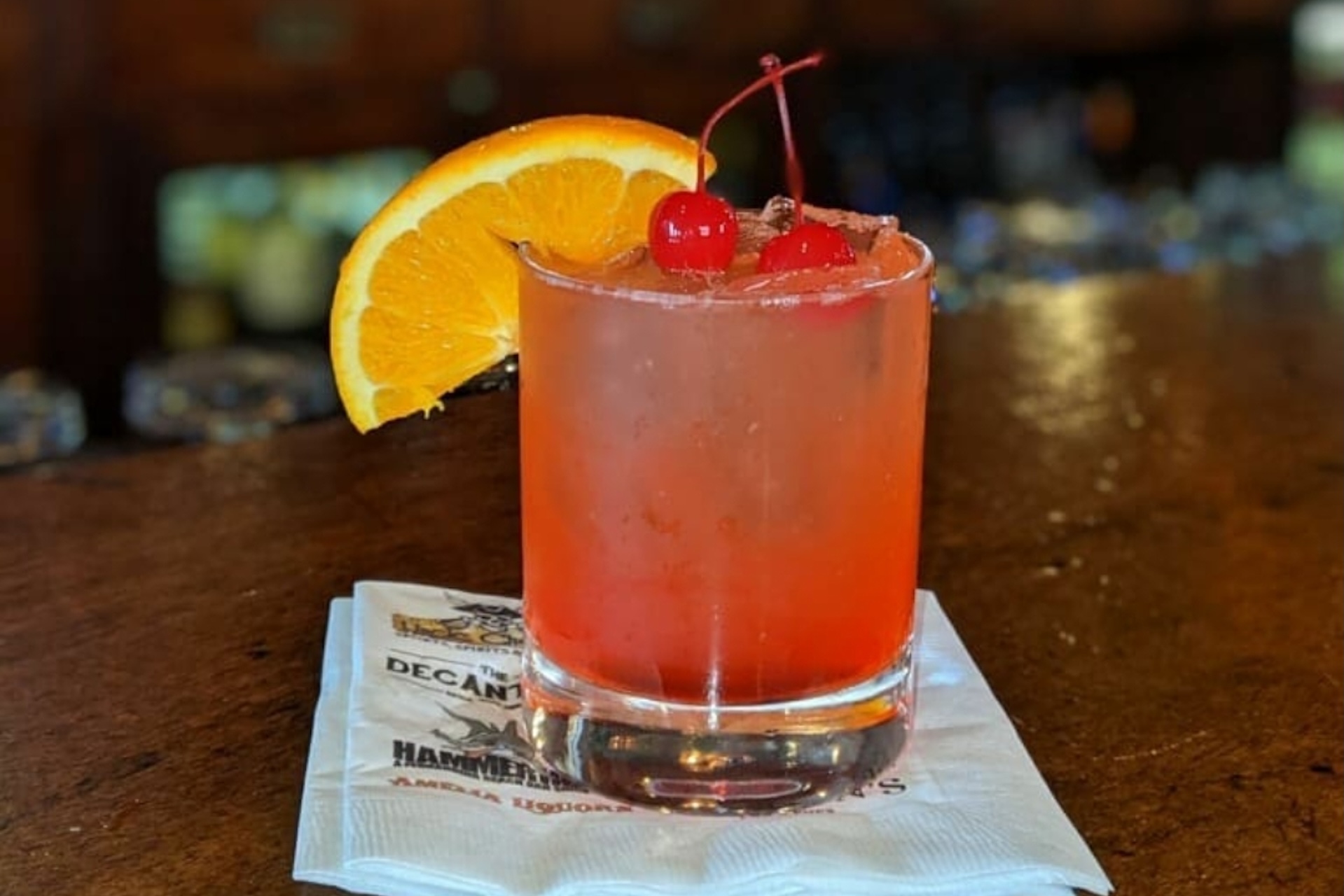 An orange-red cocktail in a glass garnished with an orange slice and two maraschino cherries, resting on a branded paper napkin.