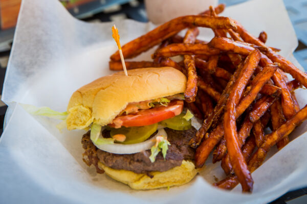A cheeseburger with lettuce, tomato, pickles, onions, and sauce on a bun, served with a side of crispy sweet potato fries on white parchment paper.