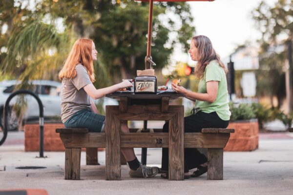 Two women sit across from each other at a wooden outdoor table, eating and talking. Trees, plants, and a car are visible in the background on a sunny day.
