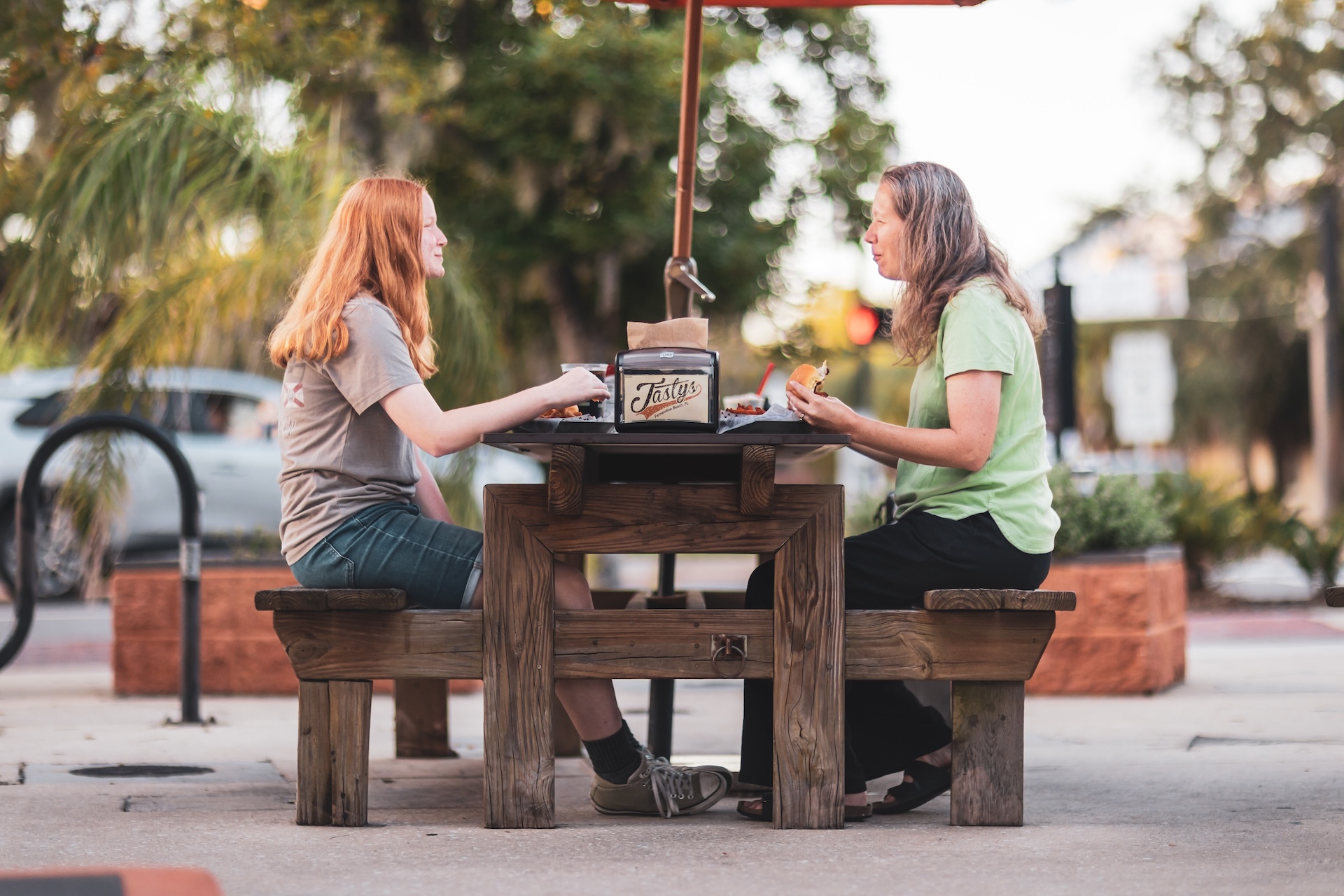 Two women sit across from each other at a wooden outdoor table, eating and talking. Trees, plants, and a car are visible in the background on a sunny day.