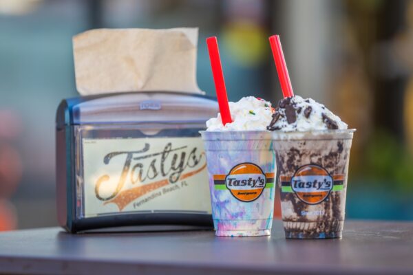 Two milkshakes topped with whipped cream and red straws sit on a table beside a napkin dispenser with the Tasty’s logo. The background is blurred.