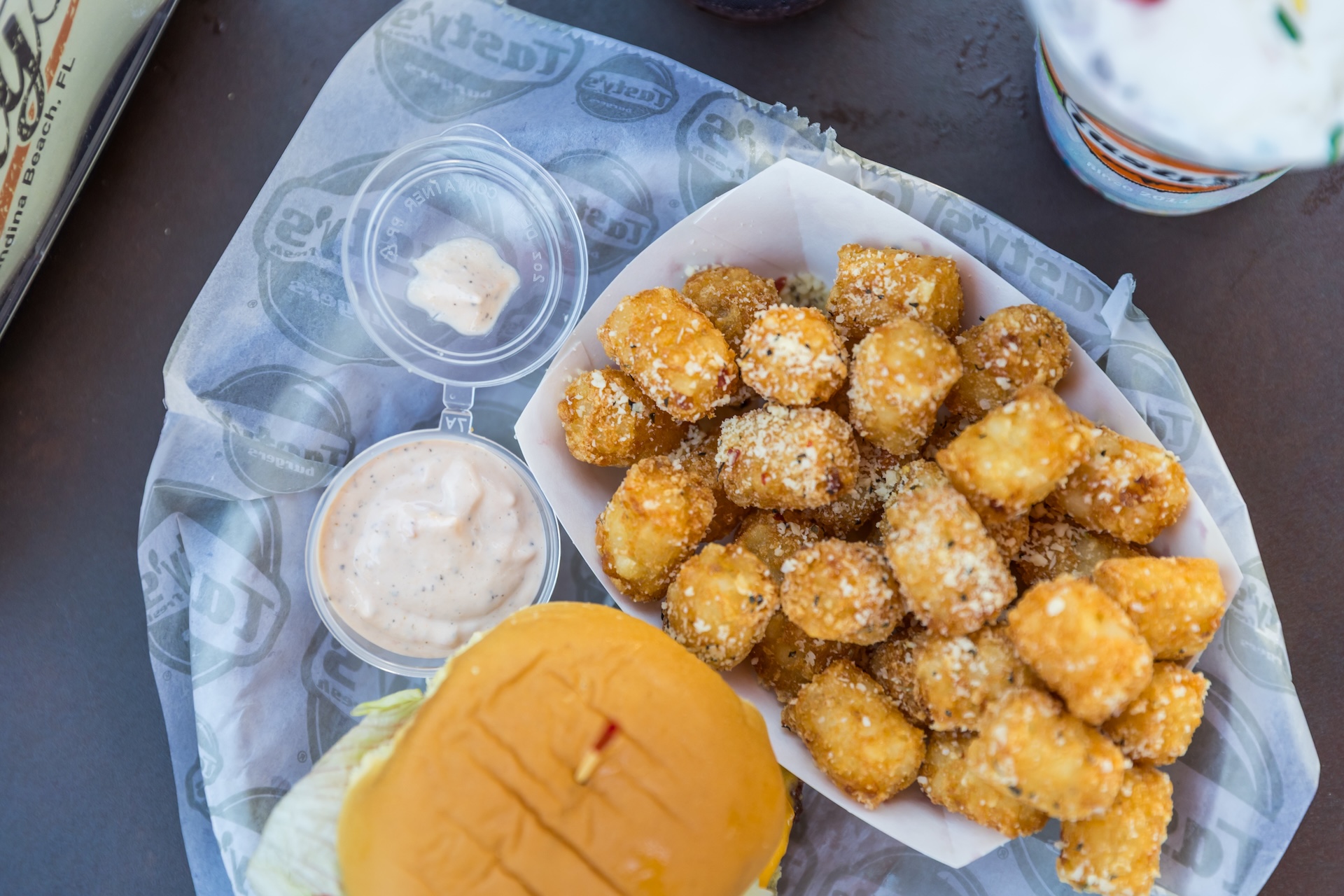 A tray with seasoned tater tots in a paper container, two cups of dipping sauce, and part of a sandwich with lettuce on a branded wrapper.