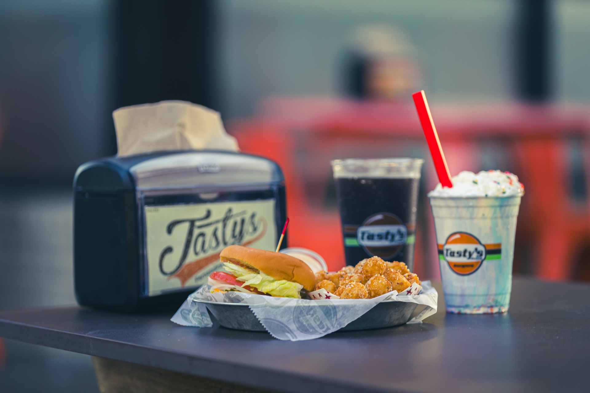A tray with a cheeseburger, tater tots, and a pickle sits next to a soft drink and a milkshake topped with whipped cream and sprinkles, with a napkin dispenser labeled “Tasty’s” in the background.