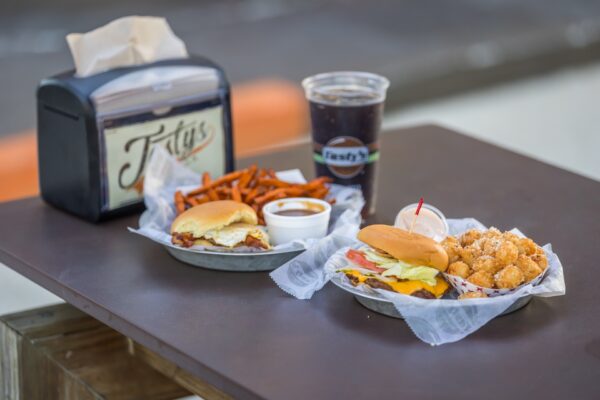 Two trays on a table, each with a burger, a side (one with sweet potato fries, one with tater tots), a small container of dipping sauce, a large cup of soda, and a napkin dispenser in the background.