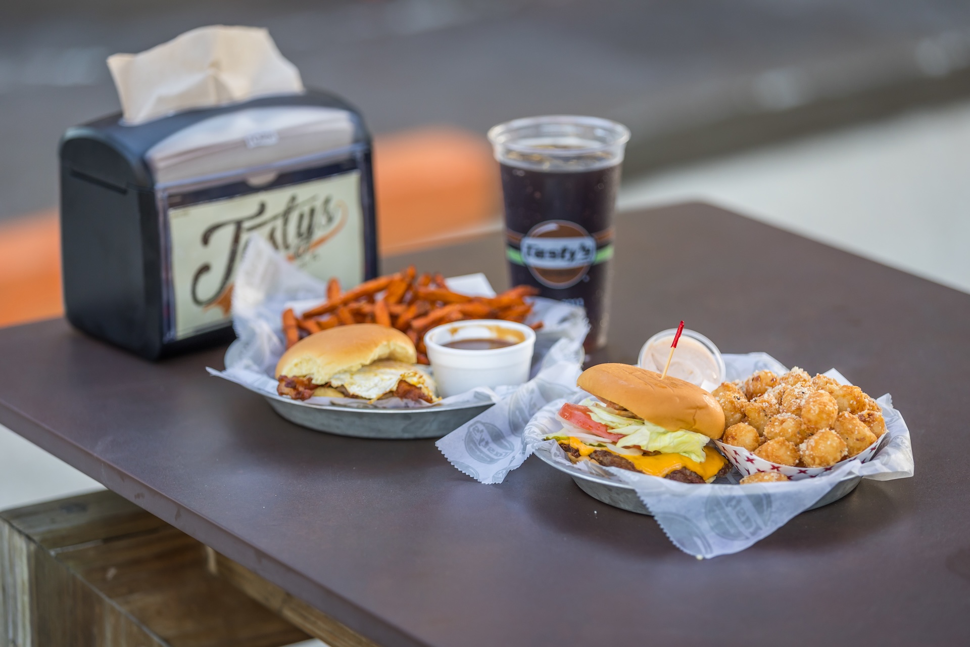 Two trays on a table, each with a burger, a side (one with sweet potato fries, one with tater tots), a small container of dipping sauce, a large cup of soda, and a napkin dispenser in the background.